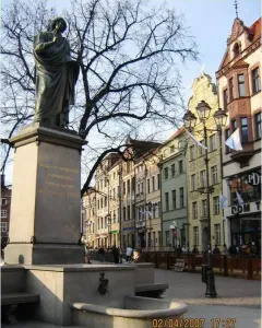 Nicolaus Copernicus Monument on a tall stone pedestal in a historic town square, with a fountain, benches, and old buildings nearby.