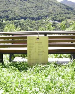A wooden bench with a hanging plaque stands in a grassy area facing a green mountainside.