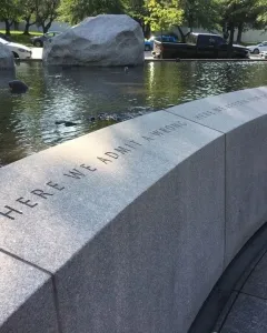 Curved stone wall of the Japanese American Memorial inscribed with “HERE WE ADMIT A WRONG,” beside a reflecting pool and boulders.
