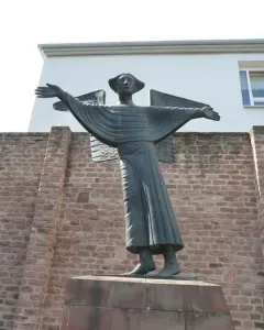 Angel of Peace statue with outstretched arms on a reddish stone pedestal, set against a brick wall and modern building.