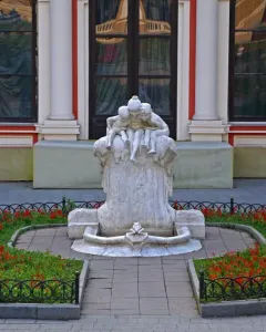 The Children and Frog sculpture on a weathered pillar, surrounded by a flowerbed with red blossoms and a low metal fence.
