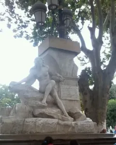Ain El Fouara Fountain with a reclining nude female statue on a rocky base and a lamppost above it, framed by a leafy tree.