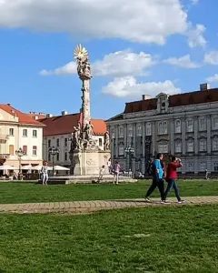 The Holy Trinity Monument stands in a sunny public square beside historic buildings and a grassy area with people walking nearby.