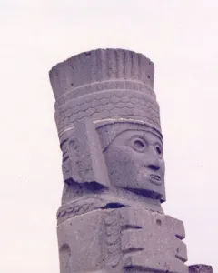 Tall stone Atlantean figure at Tula with an elaborate headdress and stylized face, seen against a bright sky.
