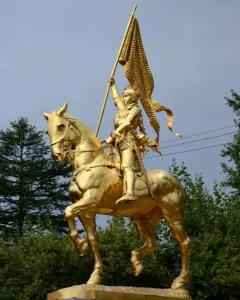 Golden equestrian statue of Joan of Arc in armor holding a flag-topped staff on a stone pedestal.