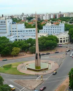 The Obelisk of Montevideo rises from a hexagonal water basin in the center of a busy traffic roundabout, with city buildings and trees around it.