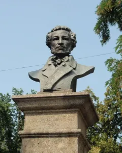 Dark bust of Pushkin on a stone pedestal outdoors, framed by trees against a blue sky.