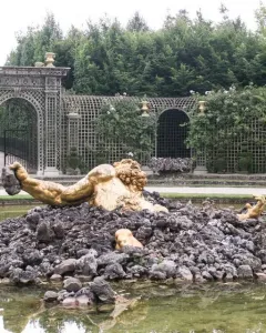 Enceladus Fountain showing a golden muscular figure rising from dark rocks in a greenish pool, with an arched trellis and greenery behind.
