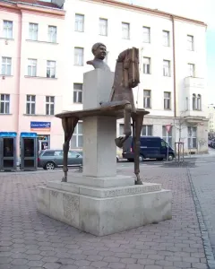 Bronze sculpture of Jaroslav Hašek with a fragmented horse on a multi-tiered stone pedestal in a paved square.
