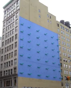 The Wall sculpture on a bright blue building facade with rows of teal projections above a beige lower section.