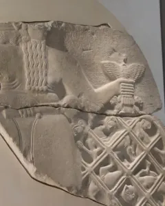 Fragmented relief from the Stele of the Vultures showing a large figure holding a staff with a bird emblem above smaller figures trapped in a net, with cuneiform text above.