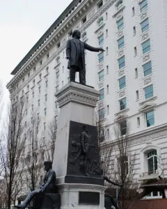 The Brigham Young Monument with a bronze figure atop a tall stone pedestal and relief sculptures at the base, with a white building and bare trees behind it.