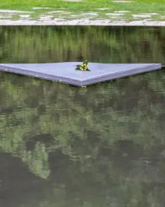 A dark triangular slab of the Memorial to the Murdered Sinti and Roma of Europe floats on water with a small cluster of flowers at its center.