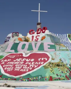 Salvation Mountain covered in colorful religious murals, with a white cross on top and large painted words including LOVE and GOD IS.