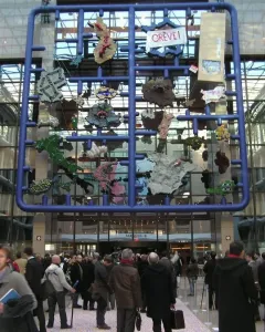 Entropa installation with a large blue grid frame and colorful attached shapes in a modern interior, with people gathered below.