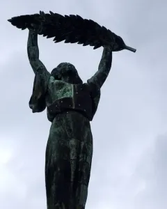 Liberty Statue bronze figure raising a leafy branch against a cloudy sky.