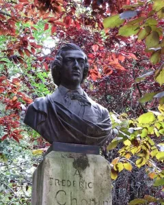 Dark metallic bust of Frédéric Chopin mounted on a weathered stone block amid red and green foliage.