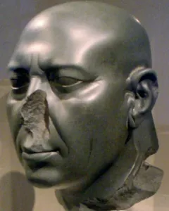 Berlin Green Head stone sculpture with a bald head, carved wrinkles, and a broken nose on a stand.