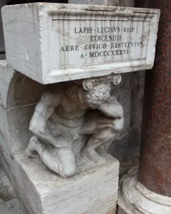 Il Gobbo di Rialto, a weathered stone figure crouching beneath an inscribed block beside a reddish column.