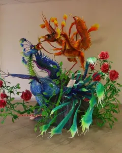 Alebrije sculpture with a blue-purple winged creature beside an orange-red bird-like figure, surrounded by green foliage and red roses.
