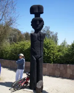 Chemamull wooden sculpture of a stylized human figure beside a bicycle on a dirt path.