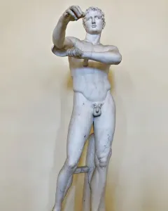 White marble Apoxyomenos statue of a nude male figure standing with one leg bearing his weight and his arms raised.