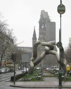 Light-grey tubular sculpture by Brigitte und Martin Matschinsky-Denninghoff in a city plaza, with a historic church ruin in the background.