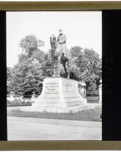 Leopold II equestrian statue on a tall stone pedestal in a park-like setting with trees and a stone balustrade behind it.