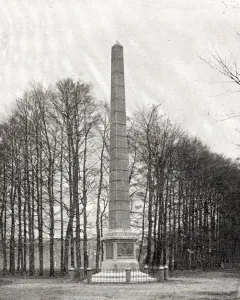 The Needle, a tall stone monument on a tiered base in a grassy clearing with bare trees behind it.