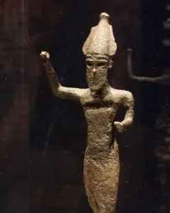 Dark metal statuette of Melqart di Sciacca with a pointed headdress and raised arm, displayed on a light base inside a case.