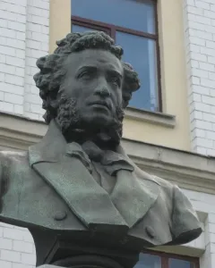 Green-patinated bronze bust of Alexander Pushkin outdoors in front of a light-colored building.