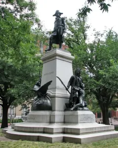 Tadeusz Kościuszko statue on a tall stone pedestal in a park, with bronze figures at the base and trees surrounding it.