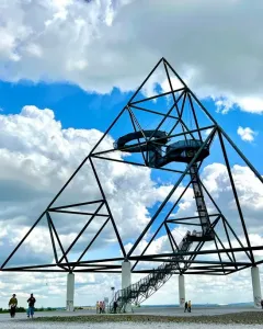 The Tetrahedron steel sculpture stands on tall white pillars with staircases and viewing platforms, with people walking nearby under a blue sky.