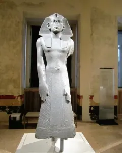 Statue of Amenemhat III standing upright in a museum gallery, wearing a striped nemes headdress and a flared kilt.