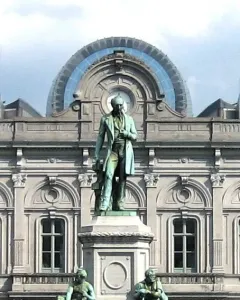 John Cockerill Monument with a standing bronze figure on an ornate pedestal and two seated figures below, in front of a grey stone building.