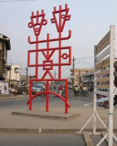 Njé Mo Yé, a bright red abstract sculpture with geometric lines and two stylized figures, stands on a concrete base beside a busy street.