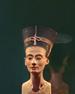 Bust of Nefertiti with a tall blue and gold crown, painted face, and patterned collar against a dark green background.