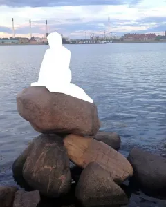 White silhouette of the Little Mermaid seated on rocks by calm water with a distant city skyline