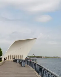 The Staten Island 9/11 Memorial postcard-shaped white monument stands on a waterfront boardwalk beside metal railings and water.
