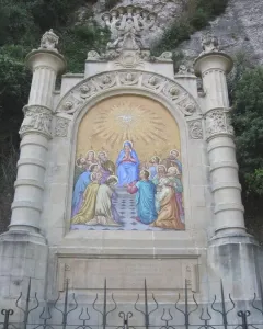 Monumental Rosary of Montserrat with a colorful Pentecost mosaic in a stone arch, seen behind a black metal fence.