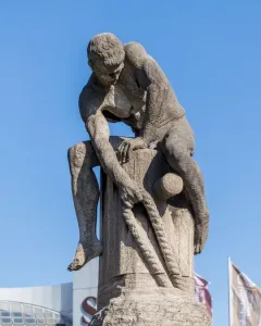Tauzieher, a weathered stone sculpture of a muscular man pulling a thick rope around a stone post.