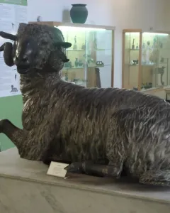 Bronze ram sculpture with curled horns resting on a stone pedestal in a museum gallery.