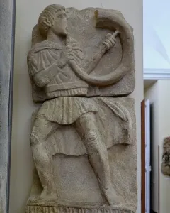Broken stone relief of the Acrobat from Osuna showing a male figure playing a horn.