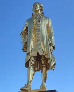 Golden statue of Alexander Hamilton in historical attire on a dark pedestal against a blue sky.
