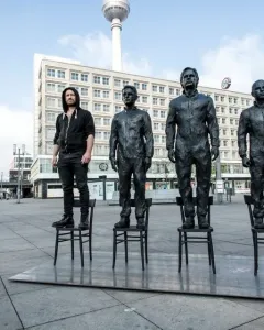Anything To Say? sculpture in Berlin, with three bronze-like statues and a live person standing on chairs in a city plaza.