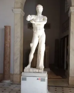 Apoxyomenos, a full-length marble statue of a nude male figure with his arms bent across his chest in a museum gallery.