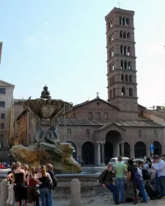 Fountain of the Tritons with sculpted figures supporting a shell-like basin, water cascading into the basin below, with a church campanile in the background.