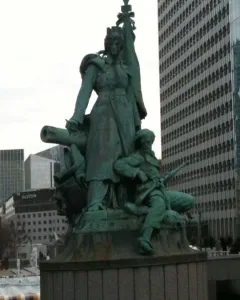 La Défense de Paris bronze monument with a crowned female figure, standard, cannon, and seated soldier in front of modern buildings