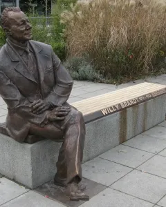 Willy Brandt Memorial bronze statue of a seated man in a suit on a bench, with tall dry grasses behind it.