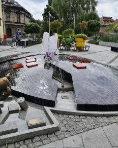 Reksio Monument beside a split egg-shaped fountain with pink-lit water in a town square.